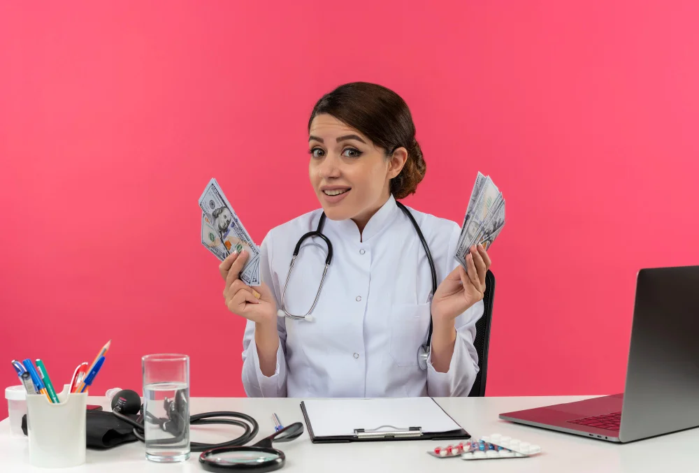 impressed-young-female-doctor-wearing-medical-robe-with-stethoscope-sitting-desk-work-computer-with-medical-tools-holding-cash-isolated-pink-wall-with-copy-space